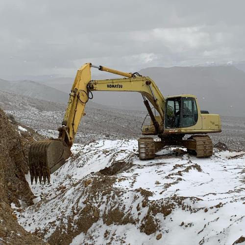 A yellow Komatsu excavator on a rocky hillside, with a cloudy sky in the background.