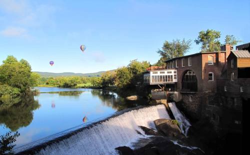 A serene river scene with a waterfall, a historic building, and hot air balloons in the clear blue sky.
