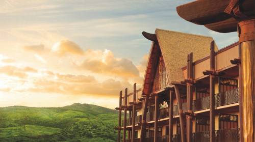 A resort building with a thatched roof against a sunset sky and green hills in the background.