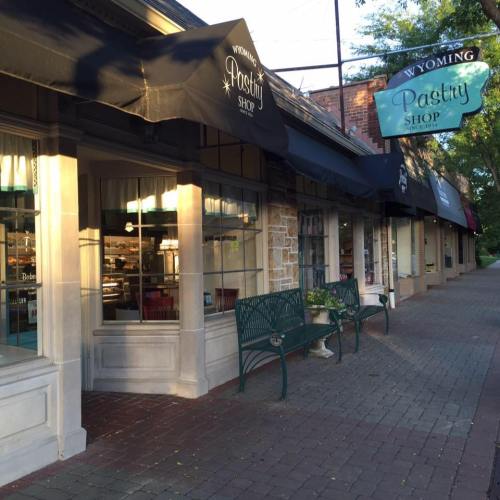 A quaint pastry shop with a welcoming entrance and outdoor seating, located on a tree-lined street.