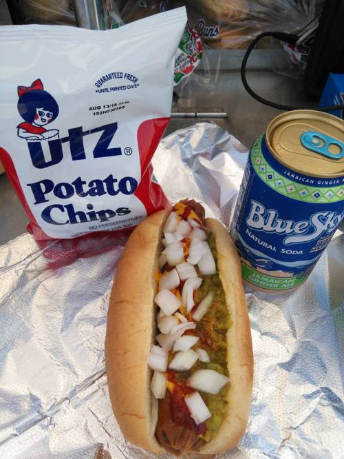 A hot dog topped with onions, mustard, and relish, alongside a bag of Utz potato chips and a can of Blue Sky soda.