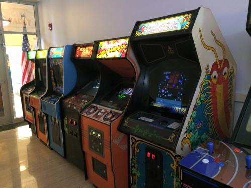 A row of vintage arcade game machines in a brightly lit room, with an American flag in the background.
