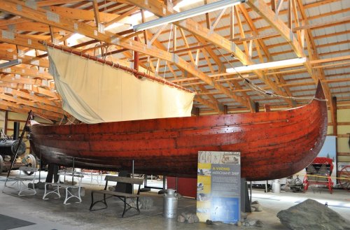 A wooden Viking ship displayed in a museum, featuring a sail and a rustic interior with wooden beams overhead.