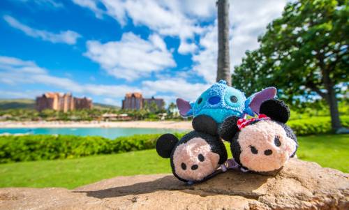 Three plush Tsum Tsum toys of Mickey, Minnie, and Stitch sit on a rock with a beach and resort in the background.