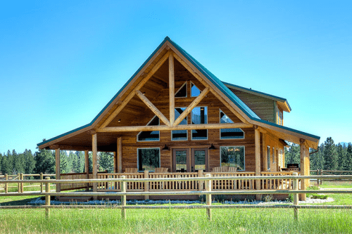 A large wooden house with a green roof, featuring a spacious porch and surrounded by a grassy field and trees.