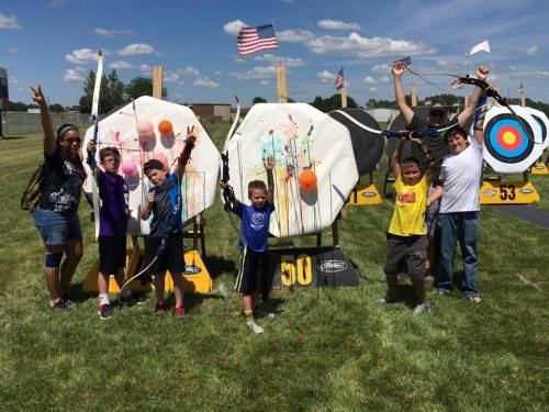 A group of children and an adult celebrate with bows and arrows in front of colorful archery targets on a sunny day.