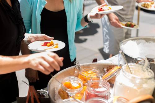 People serving themselves from a buffet with fruit and desserts, holding plates in a bright outdoor setting.