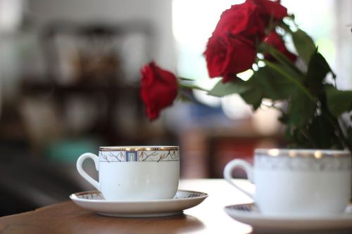 Two elegant teacups on a table, accompanied by a bouquet of red roses in the background.