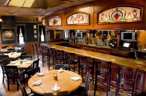 Interior of a cozy restaurant with wooden tables, chairs, and a bar featuring stained glass windows.