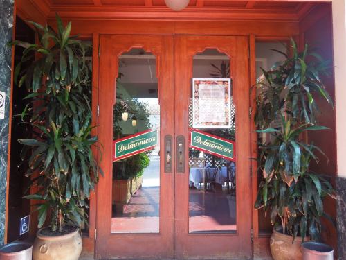 Wooden double doors with "Delmonicos" signs, flanked by potted plants, leading into a restaurant.