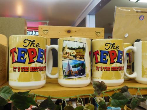 Three colorful mugs with "The TEPEE" text and images of a tepee and landscape, displayed on a wooden shelf.