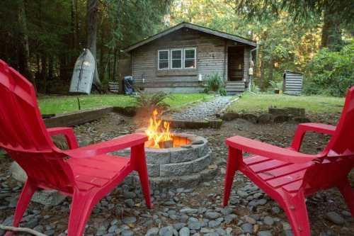A cozy cabin in the woods with a fire pit and two red chairs in the foreground, surrounded by trees and gravel.
