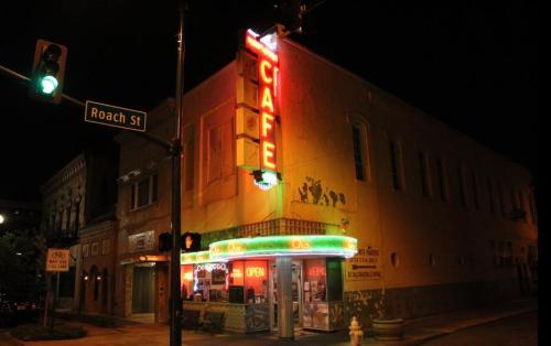 A brightly lit café at night, featuring neon signs and a corner street sign for Roach St.