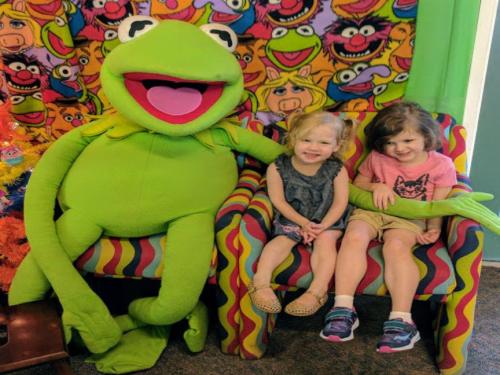 Two young girls sit on colorful chairs with a large Kermit the Frog puppet, smiling in a vibrant, playful setting.