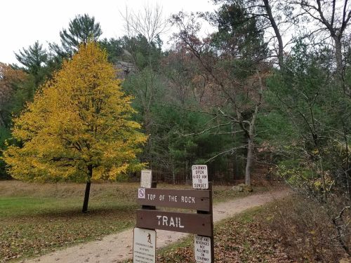 A trail sign for "Top of the Rock" surrounded by trees, with an autumn tree displaying yellow leaves nearby.