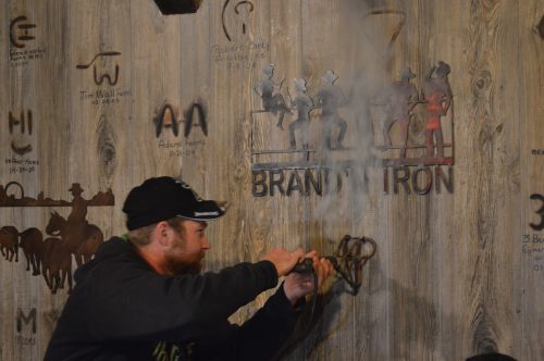 A man works with a tool near a wooden wall featuring cowboy silhouettes and names engraved on it. Smoke rises from his activity.
