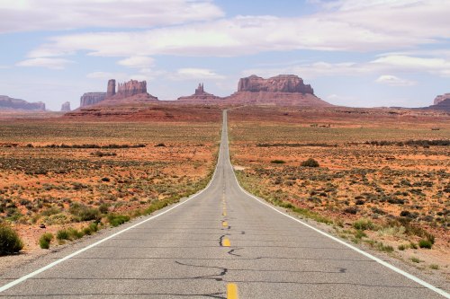 A long, empty road stretches through a desert landscape with distant rock formations under a blue sky.