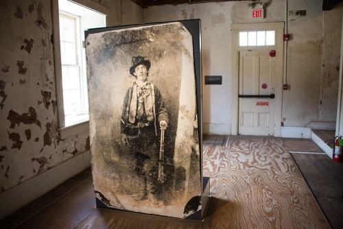 A large vintage photograph of a man in western attire displayed in a rustic room with peeling walls.