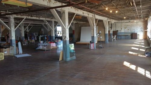 Spacious, industrial-style room with wooden floors, exposed beams, and scattered furniture and clothing items.