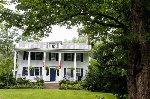 A charming white house with a porch, surrounded by greenery and trees, featuring American flags hanging from the balcony.