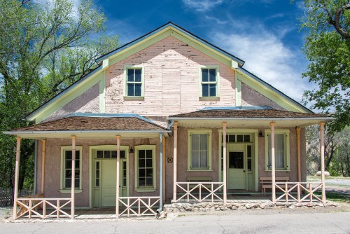 A weathered two-story house with a pink exterior, green doors, and a porch, set against a blue sky and trees.
