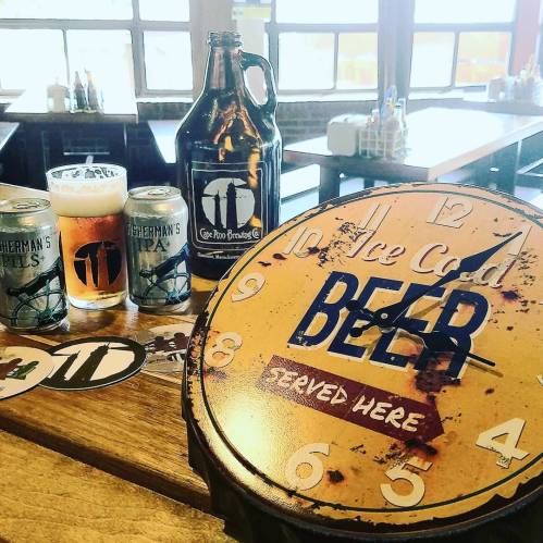 A rustic clock displays "Ice Cold Beer Served Here" next to craft beer cans and glasses on a wooden table.