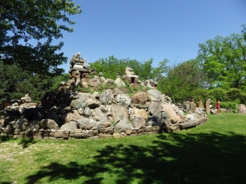 A rocky hill surrounded by greenery, with sculptures and people exploring the area under a clear blue sky.