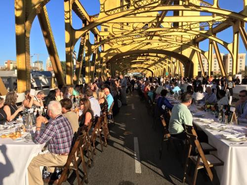 A long table set for a large outdoor dinner on a yellow bridge, with guests enjoying the meal under a clear blue sky.