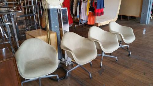 Four beige, modern chairs on wheels are lined up on a wooden floor, with clothing racks in the background.
