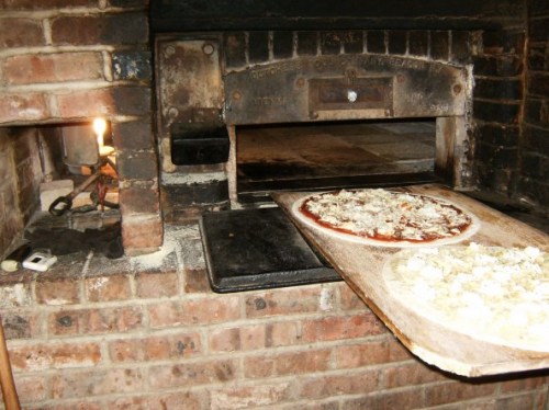 A brick pizza oven with a pizza on a wooden peel ready to be baked inside. A candle flickers nearby.