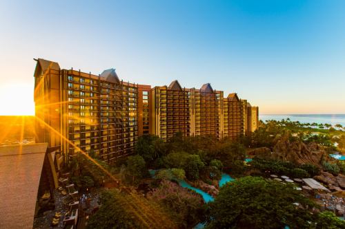 Sunset view of a large resort building surrounded by lush greenery and a pool, with the ocean in the background.