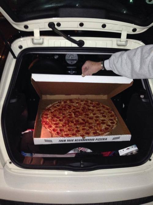 A large pepperoni pizza in an open pizza box, placed in the trunk of a car.
