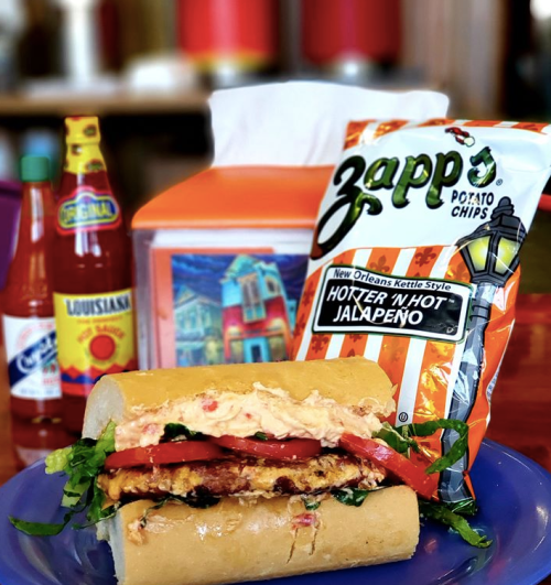 A sandwich with lettuce, tomatoes, and creamy spread, served with Zapp's potato chips and hot sauce bottles in the background.