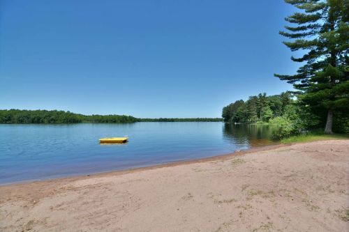 A serene lake scene with a sandy shore, a yellow float, and lush green trees under a clear blue sky.