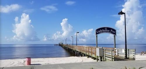 A wooden pier extends into calm waters under a blue sky with fluffy clouds, marked by a sign reading "Lighthouse Pier."