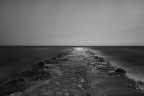 A long, rocky pier extends into a calm sea, captured in black and white under a cloudy sky.
