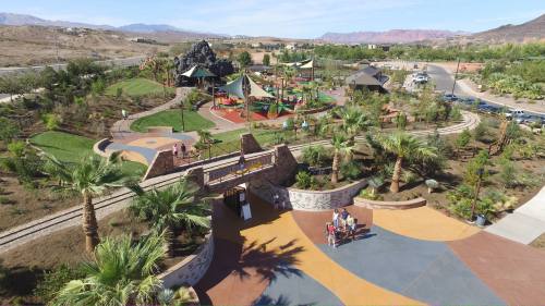 Aerial view of a colorful park with playgrounds, palm trees, and walking paths, surrounded by mountains and greenery.