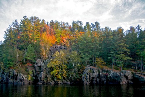 A rocky shoreline with vibrant autumn trees reflecting in calm water under a cloudy sky.