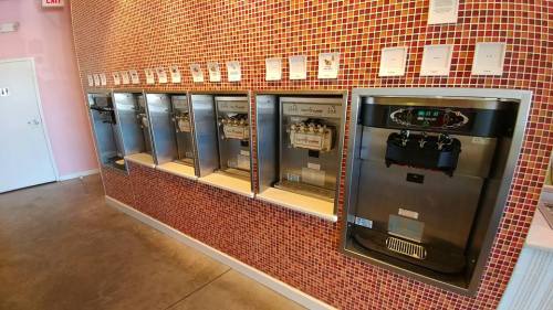 A wall with multiple frozen yogurt machines, each with a digital display, set against a colorful tiled background.