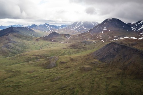Aerial view of rugged mountains and rolling green hills under a cloudy sky, with patches of snow on the peaks.