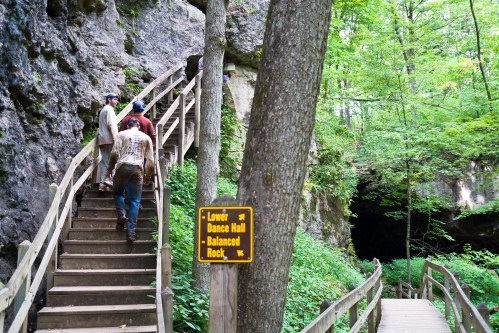 Hikers ascend wooden stairs in a lush forest, with a sign for "Lower Dance Hall" and "Balanced Rock" nearby.