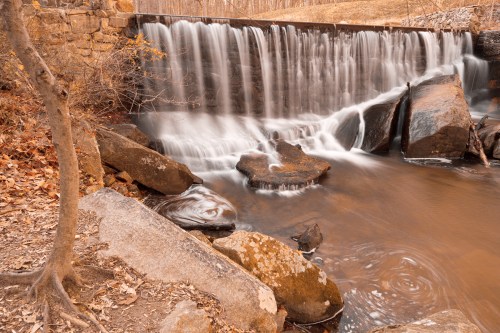 A serene waterfall cascades over rocks, surrounded by autumn leaves and a tranquil stream.