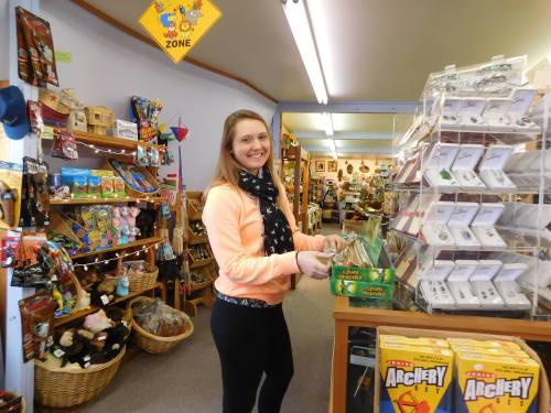 A smiling woman stands at a shop counter, surrounded by colorful merchandise and crafts in a cozy store.