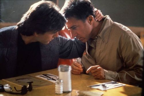 Two men sit closely at a diner table, engaged in a serious conversation, with a salt shaker and utensils visible.