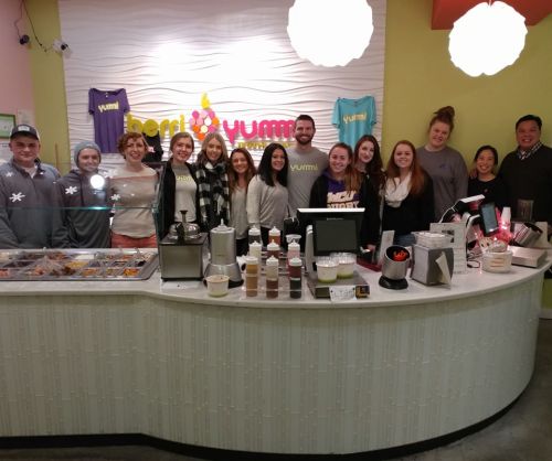 A group of smiling staff members stands behind a colorful counter at a frozen yogurt shop, with toppings and machines visible.