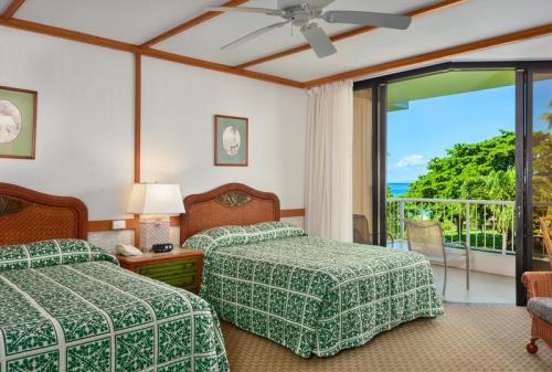 A bright hotel room with two beds, tropical-patterned bedding, a lamp, and a balcony overlooking greenery and ocean views.