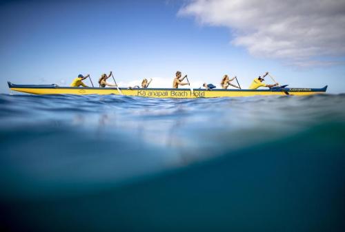 A group of people paddling in a yellow canoe on clear water, with a beach hotel in the background.