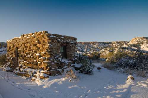 A stone building covered in snow, surrounded by a winter landscape of hills and sparse vegetation under a clear blue sky.