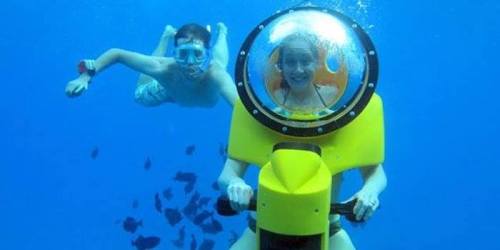 A person in a yellow underwater scooter smiles while another swimmer explores nearby in clear blue water.
