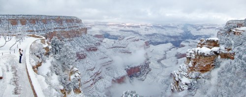 A panoramic view of a snowy Grand Canyon, with mist rising from the canyon and a person taking photos on the edge.